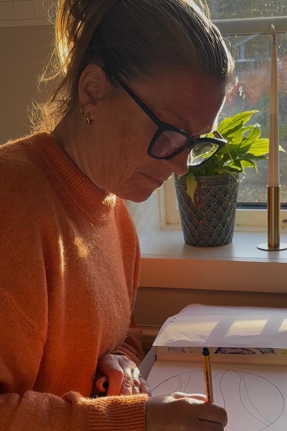 Person sitter framför fönstret i kvällsol och ritar